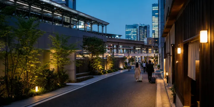 Travelers with suitcases walk through a calm Osaka-style urban hotel district at dusk.